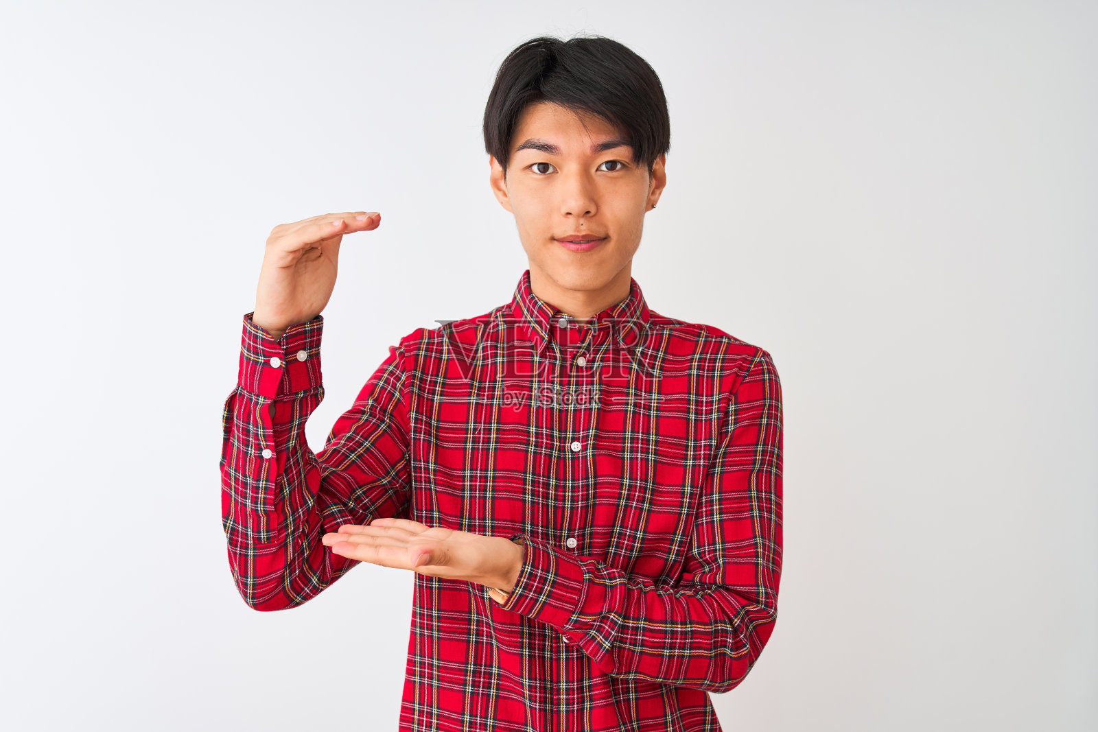 一名年轻的中国男子穿着休闲的红色衬衫站在孤立的白色背景下打手势，双手展示着大尺寸的标志，测量符号。看着镜头微笑。测量的概念。照片摄影图片