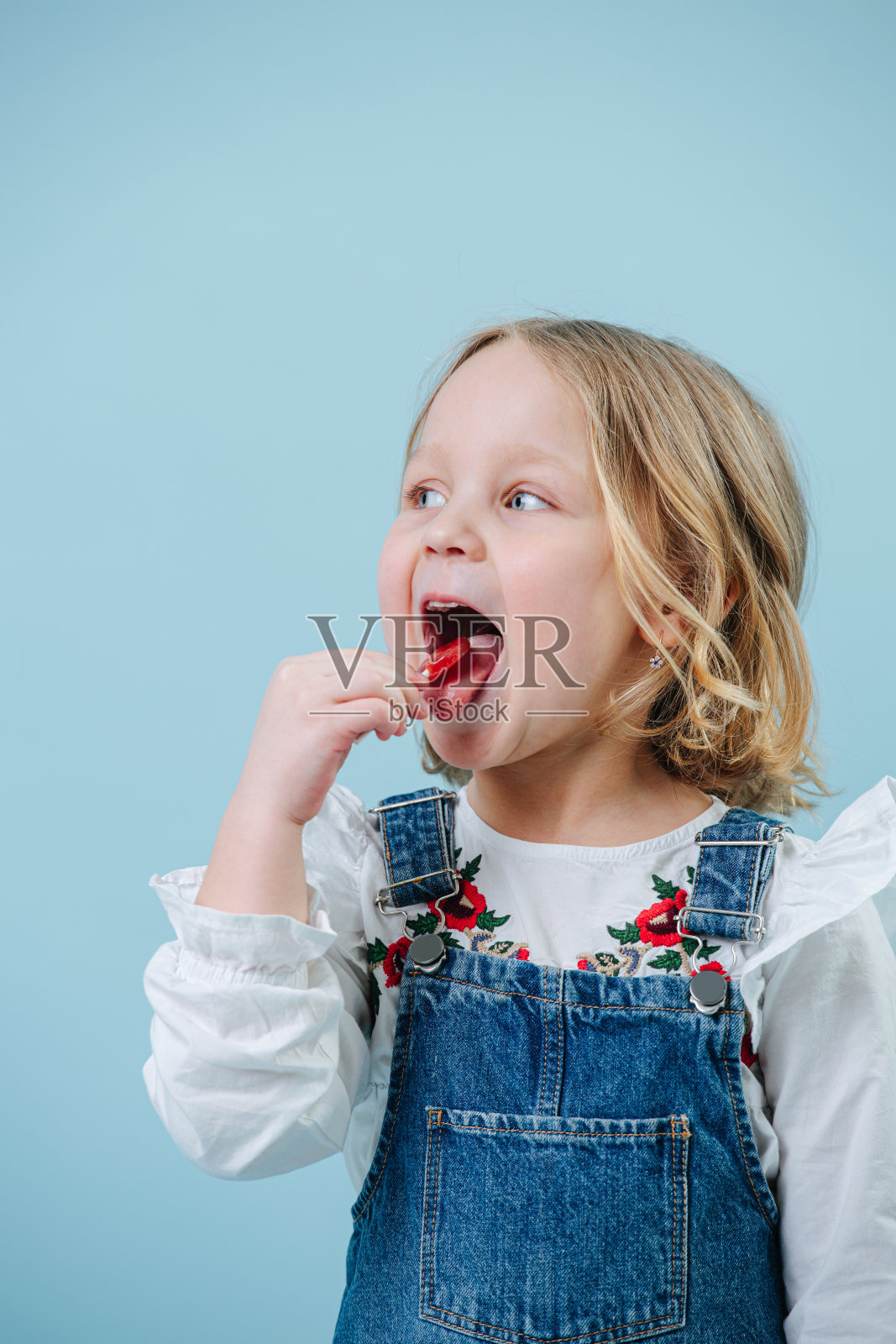 快乐的金发小女孩穿着牛仔裤，喜欢吃棒棒糖而不是蓝色照片摄影图片