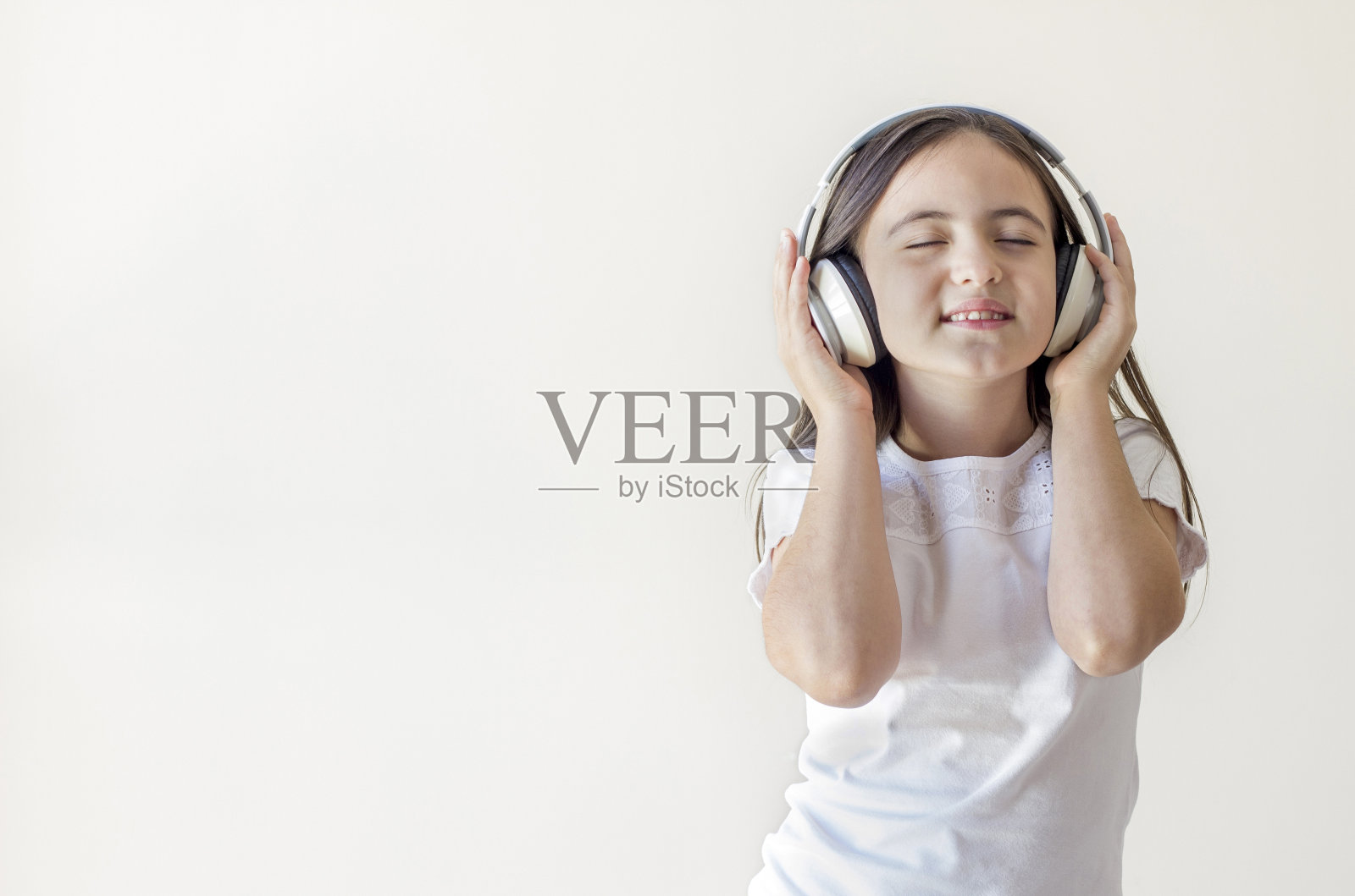 一个穿着白色衬衫的黑发小女孩的肖像，戴着大大的无线耳机，听着音乐，闭着眼睛唱歌跳舞，没有人在家照片摄影图片