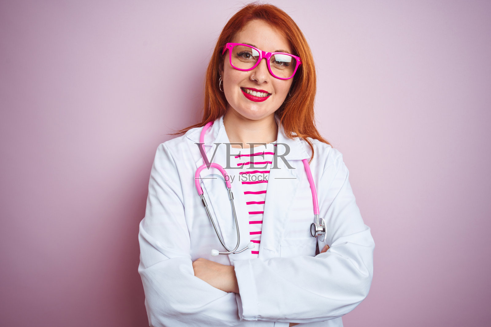 年轻的红发女医生使用听诊器站在孤立的粉红色背景快乐的脸微笑，双臂交叉看着相机。积极的人。照片摄影图片
