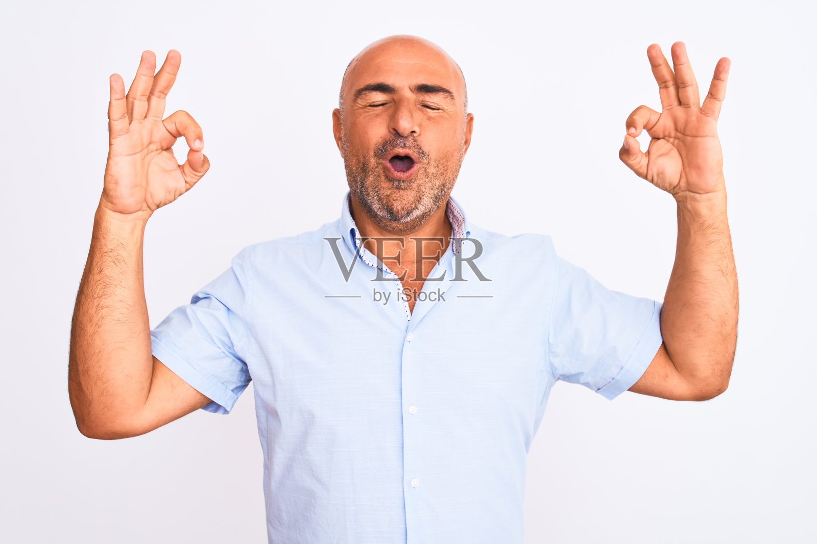 穿着休闲衬衫的中年帅气男人，在白色背景前闭着眼睛放松微笑，用手指做冥想手势，瑜伽概念照片摄影图片