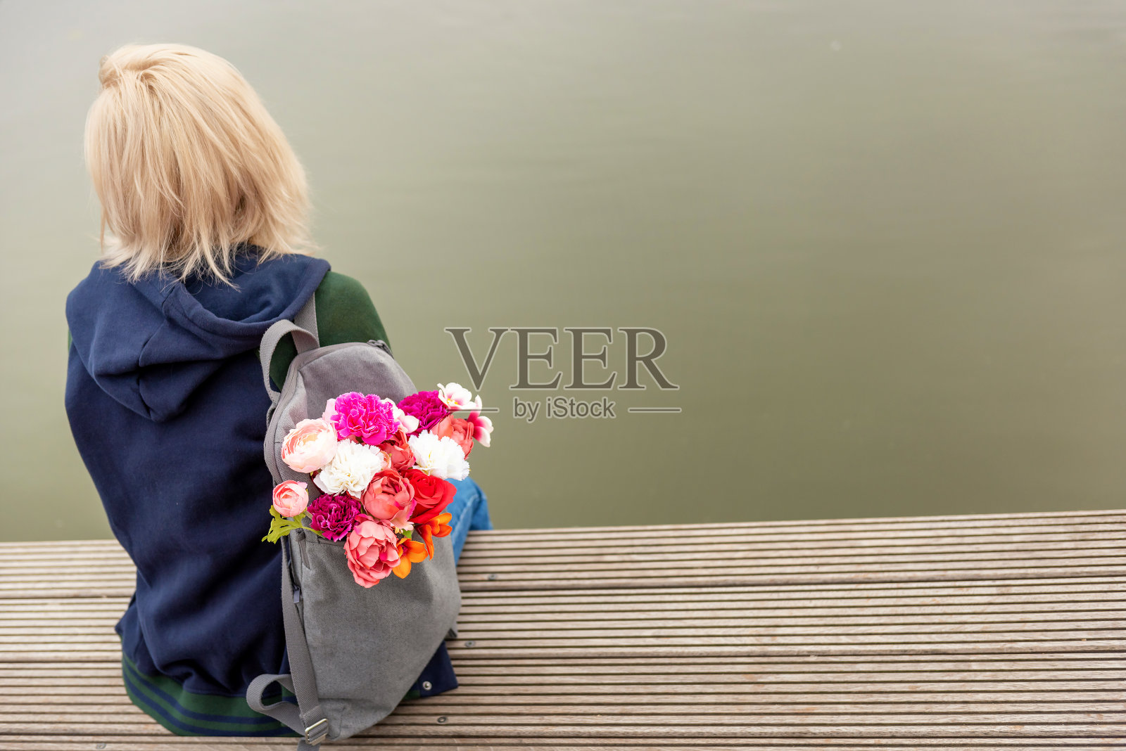 年轻金发梦幻女子在河边拿着一束鲜花，灰色背包的背影照片，浪漫氛围照片摄影图片