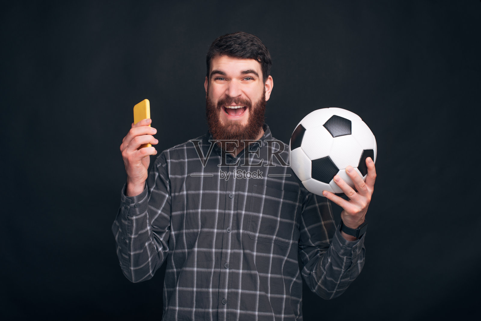 一个长着胡子的快乐的年轻人正拿着手机和一个足球靠近黑色的墙壁。照片摄影图片