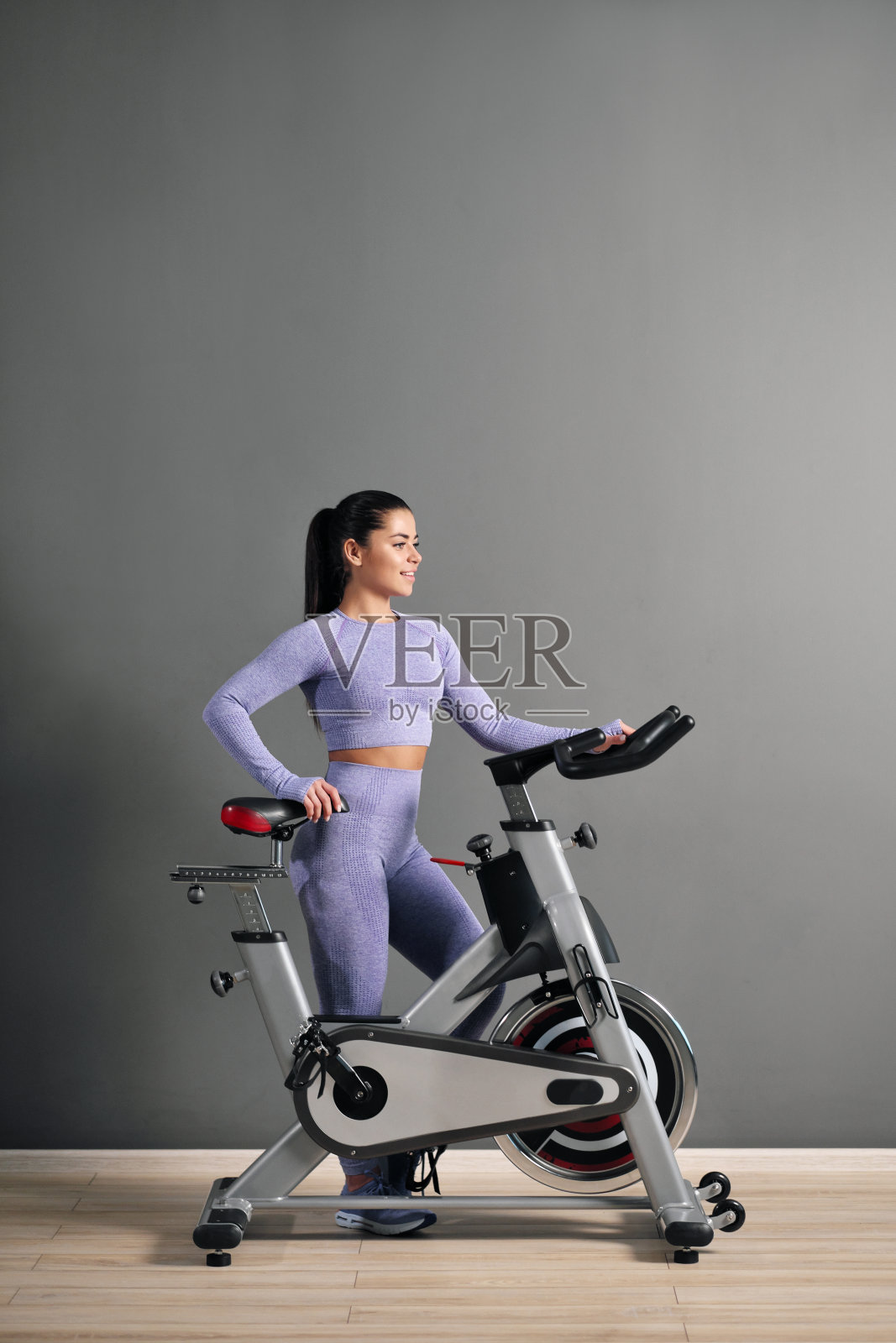 一个漂亮的运动年轻女子身穿运动服，骑着自行车在体育馆的灰色墙壁上。照片摄影图片