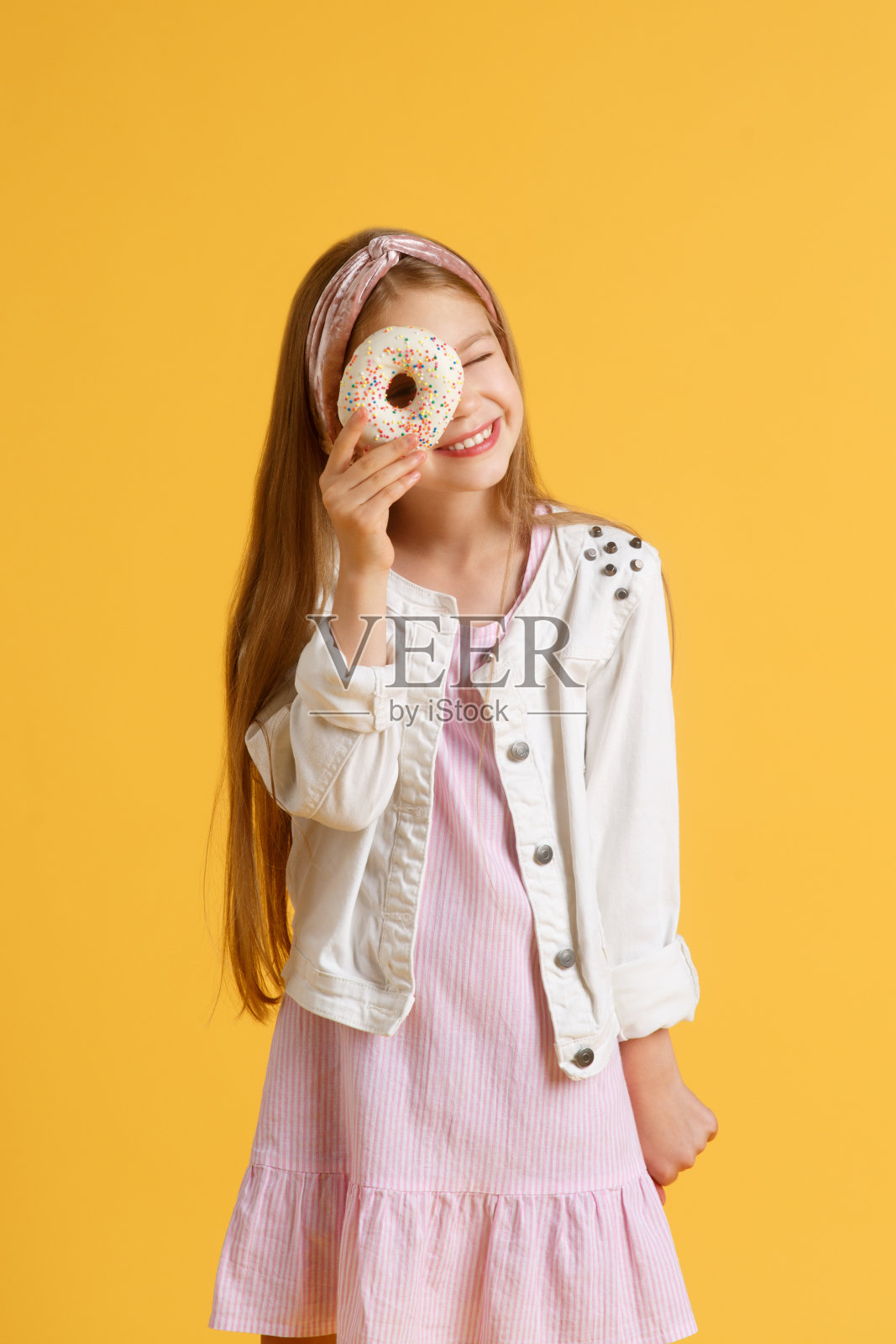 疯狂快乐的金发女孩戴着生日帽微笑，玩得很开心，看着她眼睛上的两个红色甜甜圈。糖果。黄色工作室背景照片摄影图片