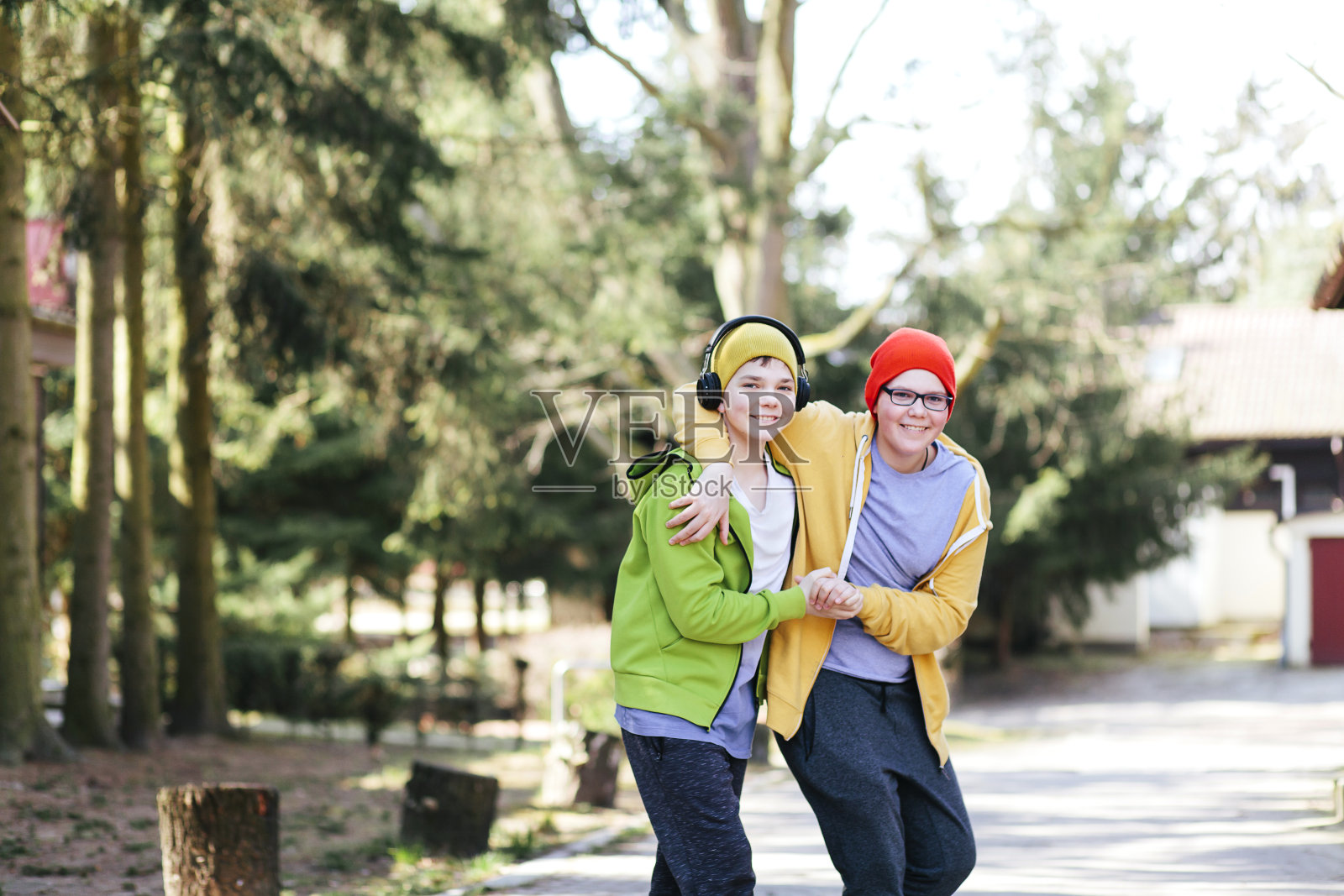 十几岁的男孩，兄弟，粗糙的住房，而站在阳光灿烂的公园，穿着绿色，黄色的帽衫，红色的帽子，耳机。照片摄影图片
