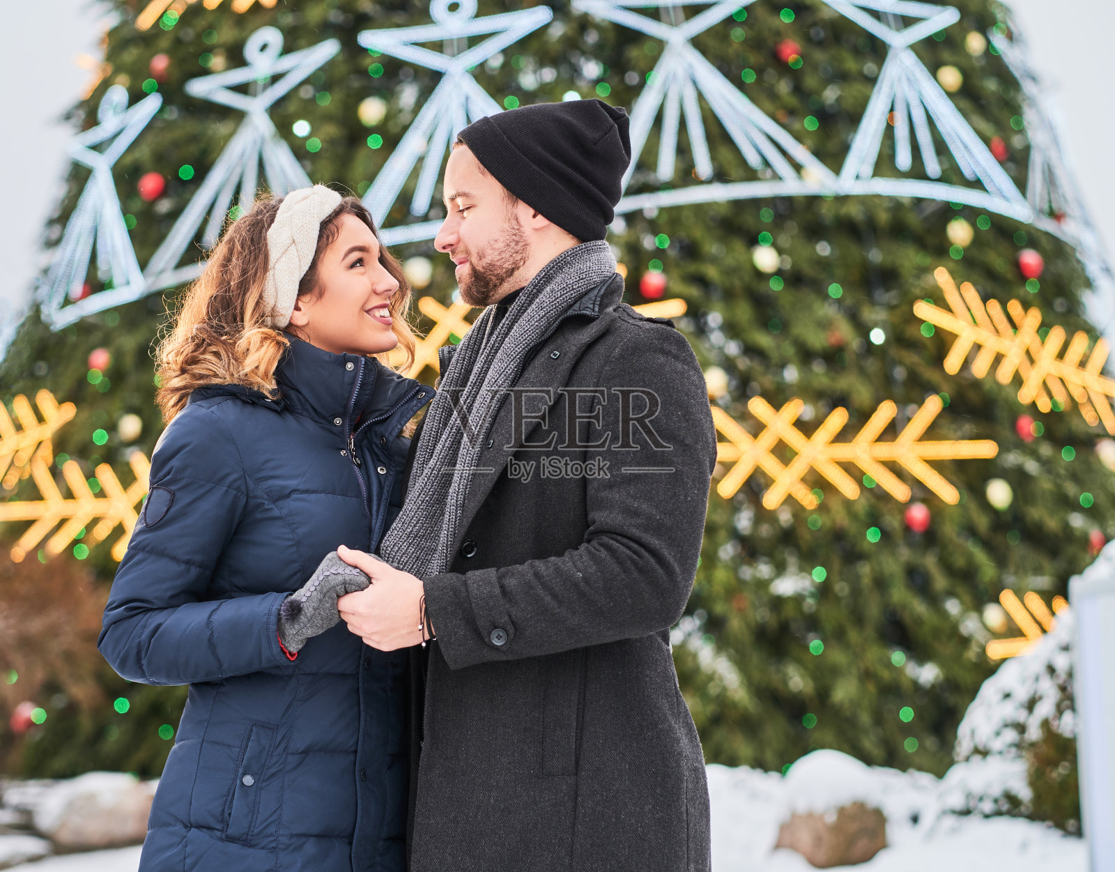 年轻夫妇穿着暖和的衣服，手牵着手，看着对方，站在城市的圣诞树旁，享受一起度过的时光。照片摄影图片