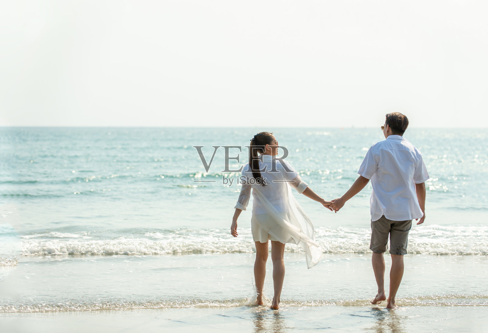 快乐的年轻亚洲夫妇在夏季牵手走在沙滩上的后视图。健康的人们在浪漫的暑假中享受放松的生活方式。照片摄影图片