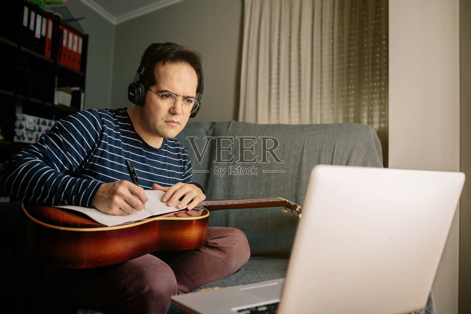 一个男孩在网上学习如何弹古典吉他。他坐在家里的沙发上，一边在笔记本电脑前弹吉他，一边听着课照片摄影图片