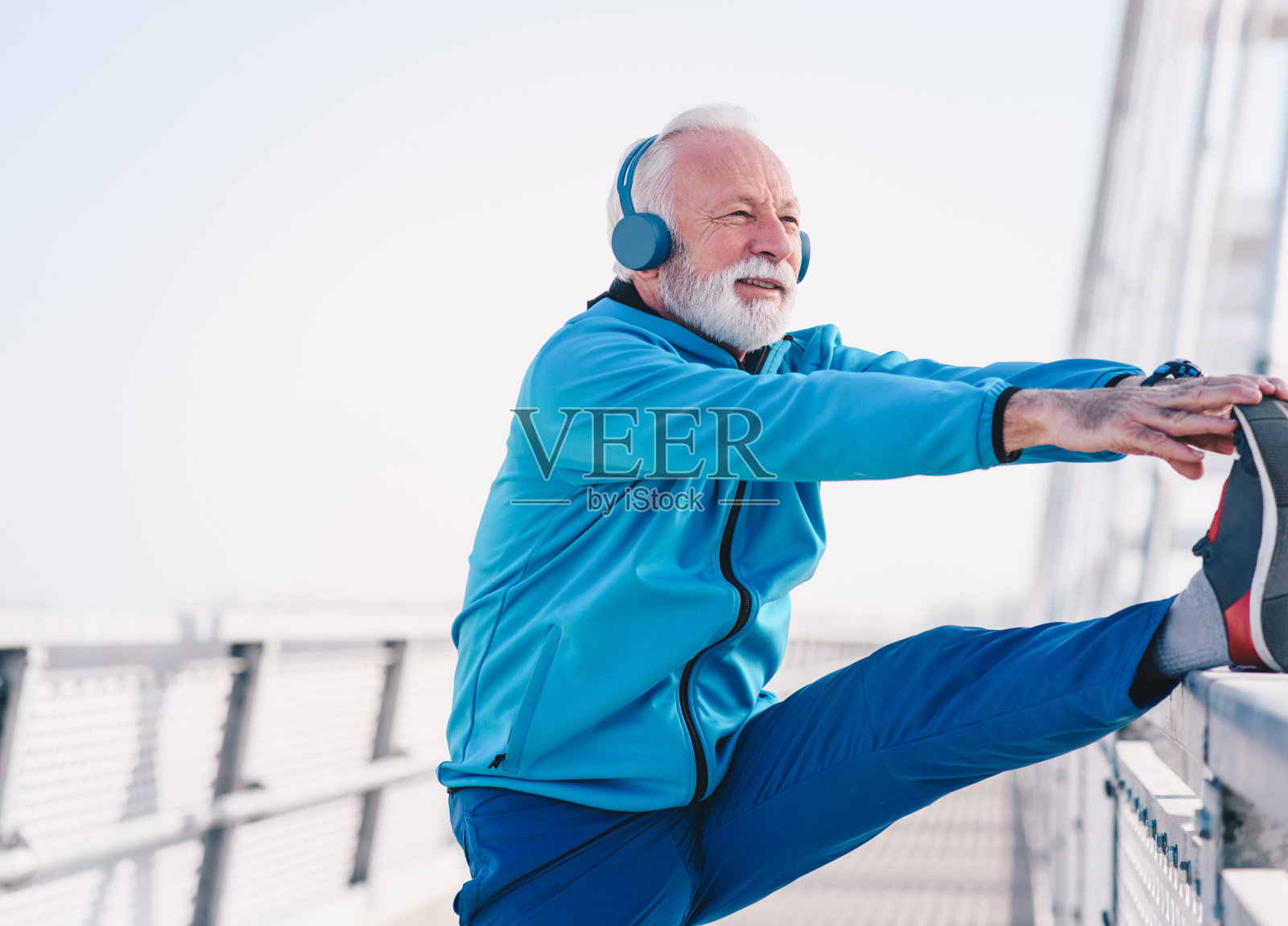 一位资深运动员在桥上的人行道上伸展他的双腿。他在横木上抬起一条腿的脚趾，拉着腿伸展。他戴着耳机。照片摄影图片