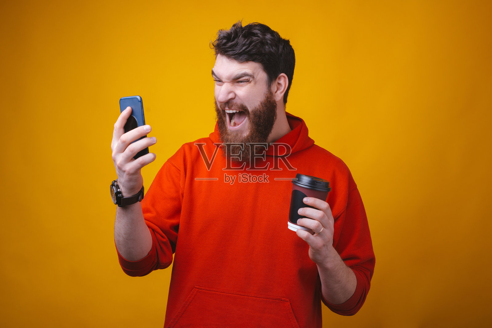 胡子疯男人一边拿着咖啡杯一边对着手机大喊大叫。照片摄影图片