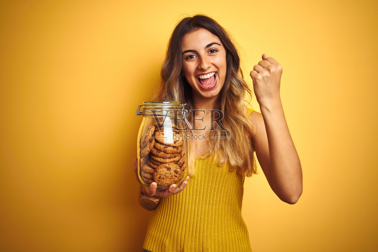 年轻漂亮的女人拿着一罐饼干，在黄色背景前尖叫，自豪地庆祝胜利和成功，非常兴奋照片摄影图片