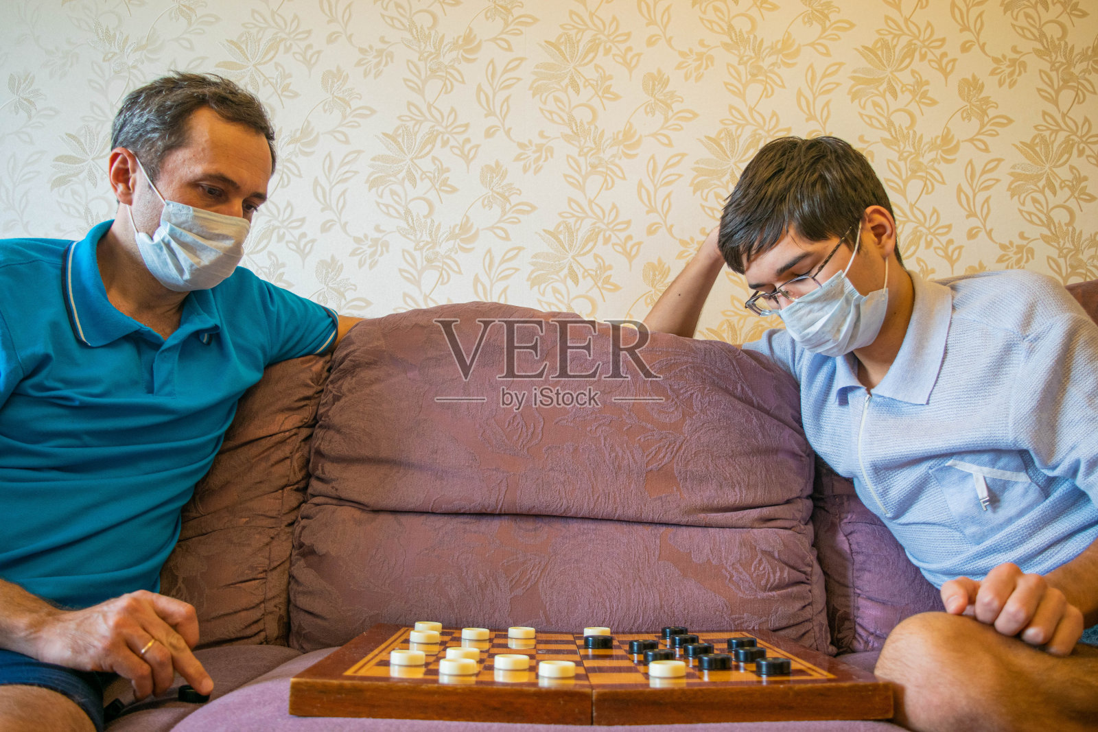 爸爸和十几岁的儿子戴着防护面具下跳棋。照片摄影图片