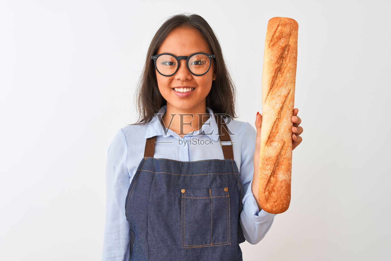 年轻的中国面包师戴着眼镜，在孤立的白色背景下拿着面包，开心地站着，自信地笑着露出牙齿照片摄影图片
