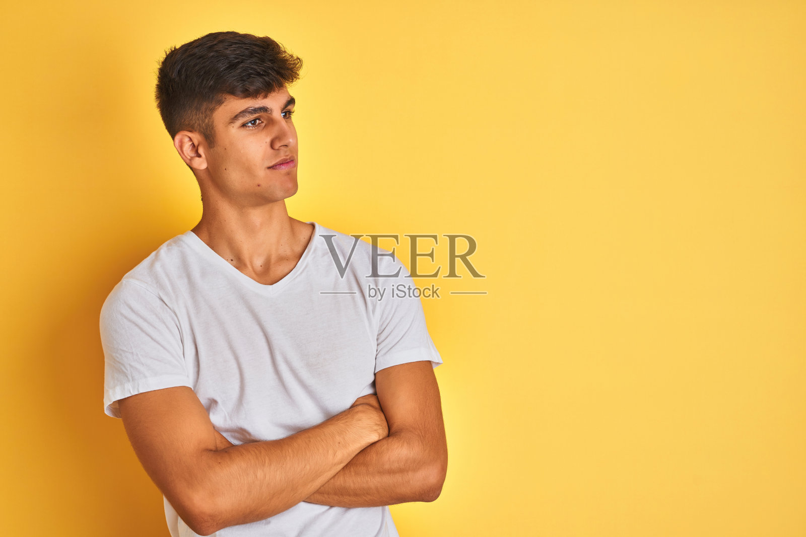 身穿白色T恤的年轻印度男子在黄色背景前叉着胳膊看向一侧，显得自信照片摄影图片