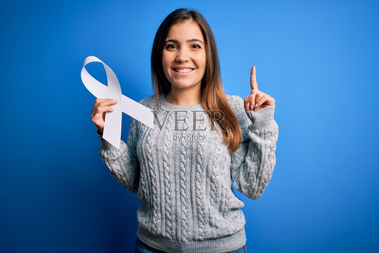 年轻金发女子拿着癌症意识和停止妇女暴力白色丝带，惊讶地有了一个主意或问题，用快乐的表情指着手指，比出第一照片摄影图片