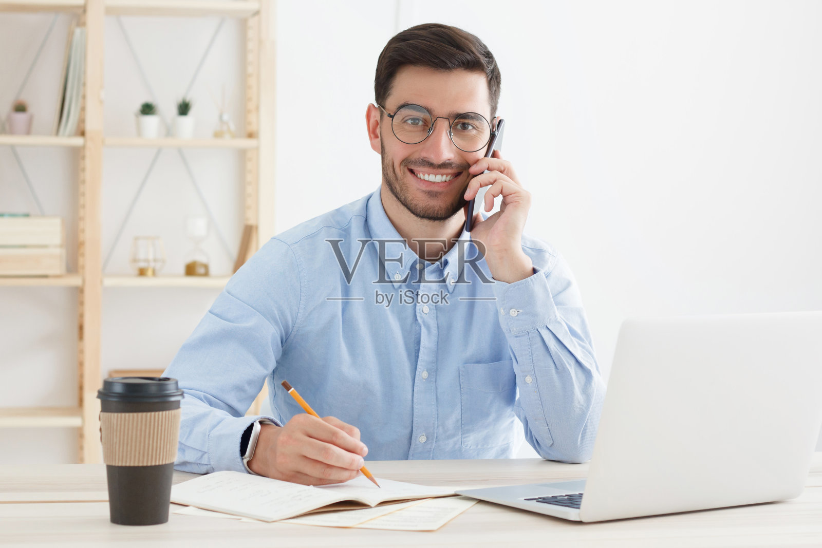 年轻的商人在办公室工作，穿着蓝色衬衫，戴着眼镜，接电话，做笔记，正面微笑照片摄影图片