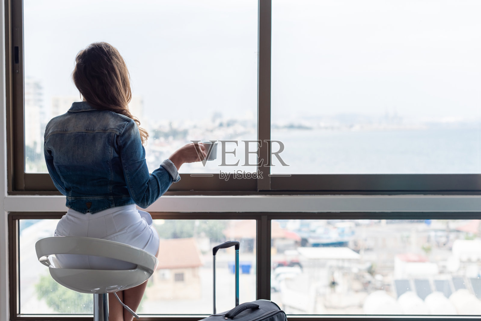 美丽的独自黑发女人穿着牛仔夹克和短裤坐在高椅子上，看着她公寓的巨大窗户。女性提着行李箱准备上路。照片摄影图片