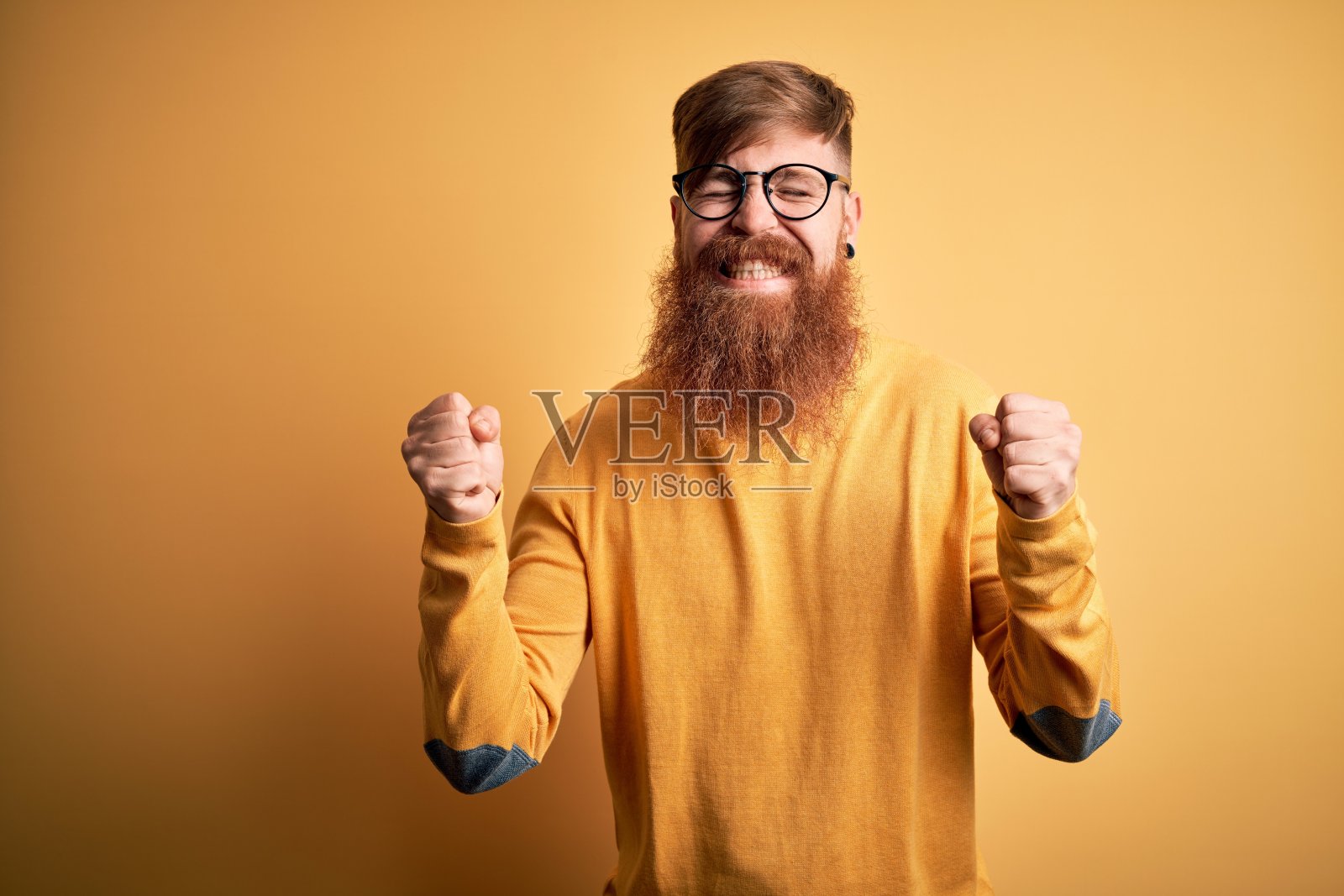 英俊的爱尔兰红发男子，留着胡子，戴着眼镜在黄色孤立的背景兴奋的成功，举起手臂，闭上眼睛庆祝胜利微笑。赢家的概念。照片摄影图片
