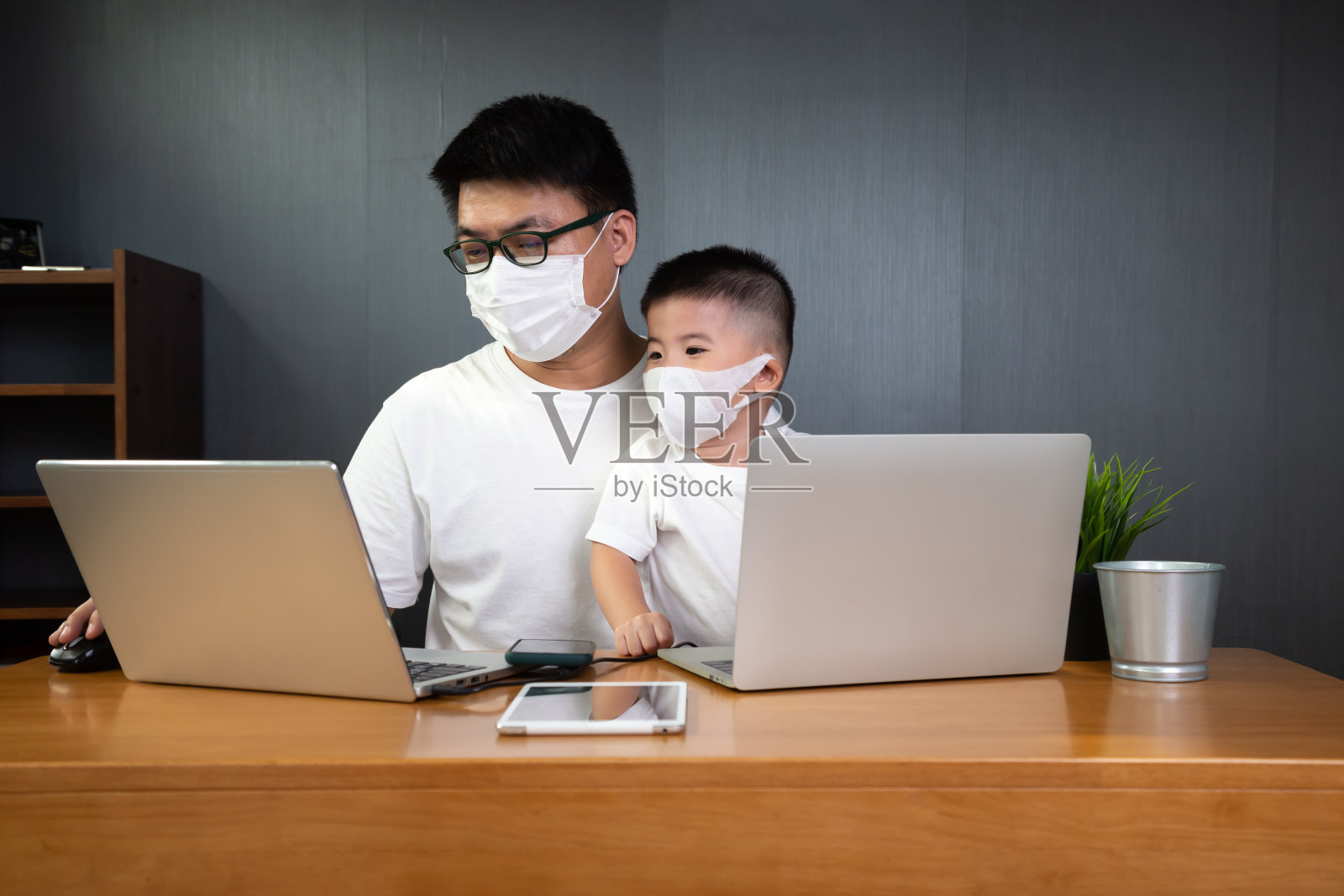 一名亚洲男子，他支持在笔记本电脑上与儿子远程工作，并戴上防护口罩，预防新冠病毒。在家工作概念照片摄影图片
