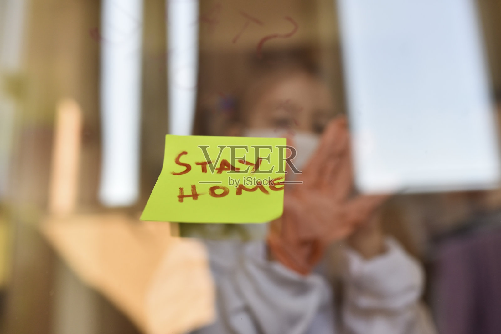 有一张脸的女孩站在窗前，向外看，并显示出“呆在家里”的信息照片摄影图片