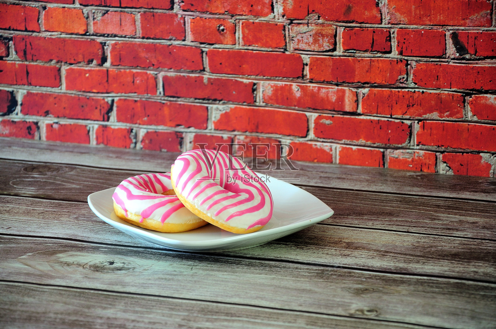 靠砖墙的木桌上放着一个白色盘子，上面放着两个草莓色的甜甜圈。照片摄影图片