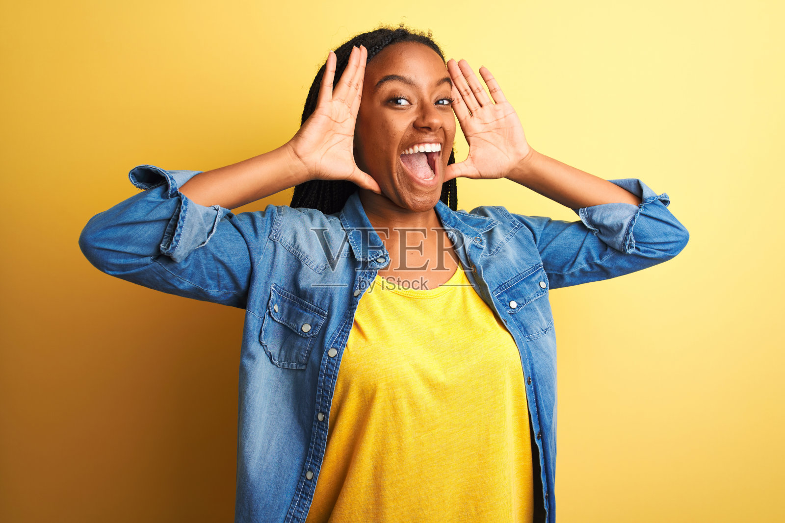 年轻的非洲裔美国妇女穿着牛仔衬衫站在孤立的黄色背景微笑愉快地玩peek a boo与手露出脸。惊讶和退出照片摄影图片