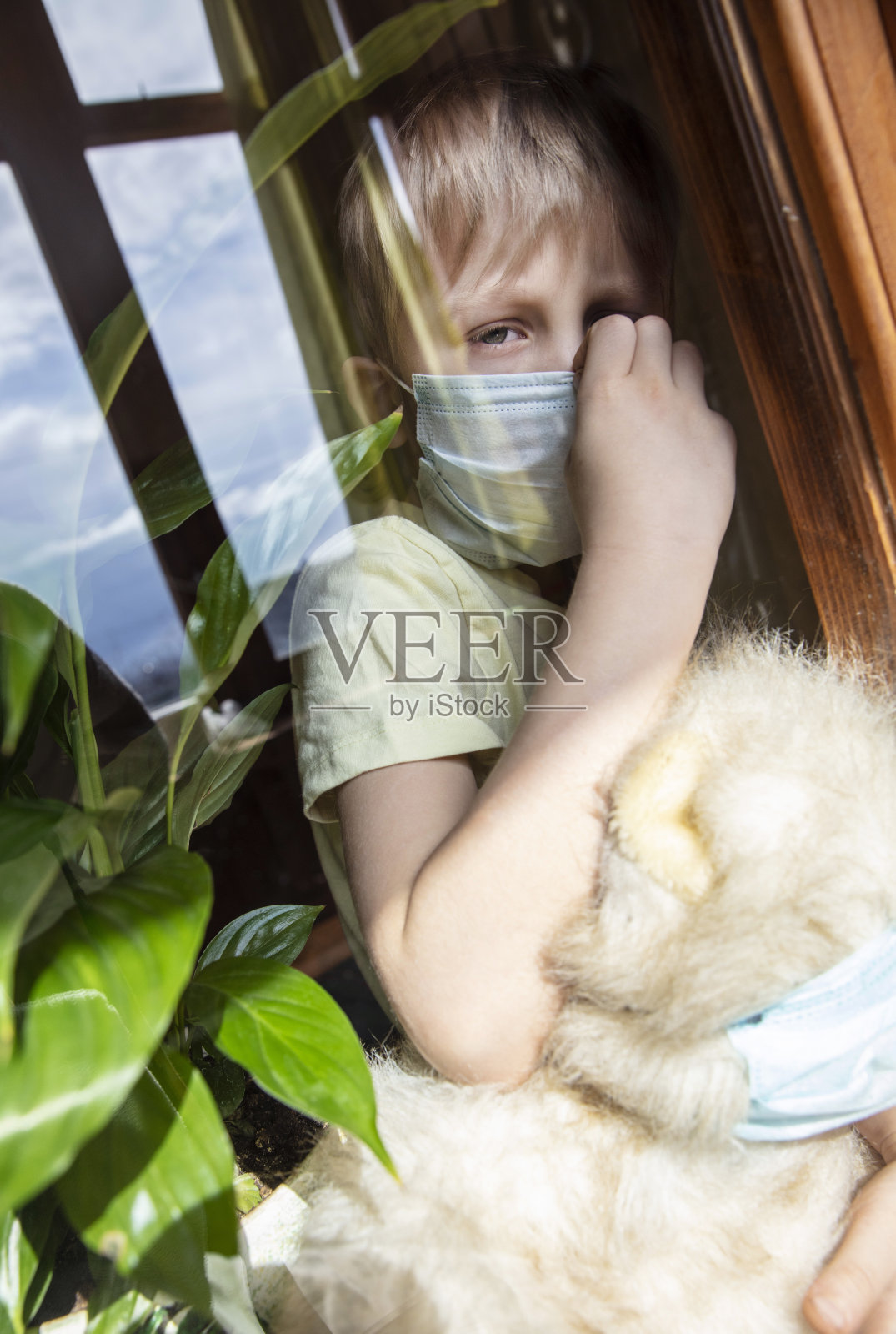 一个男孩带着他生病的泰迪熊，戴着医用防护口罩从窗户往外看。预防流行病。照片摄影图片