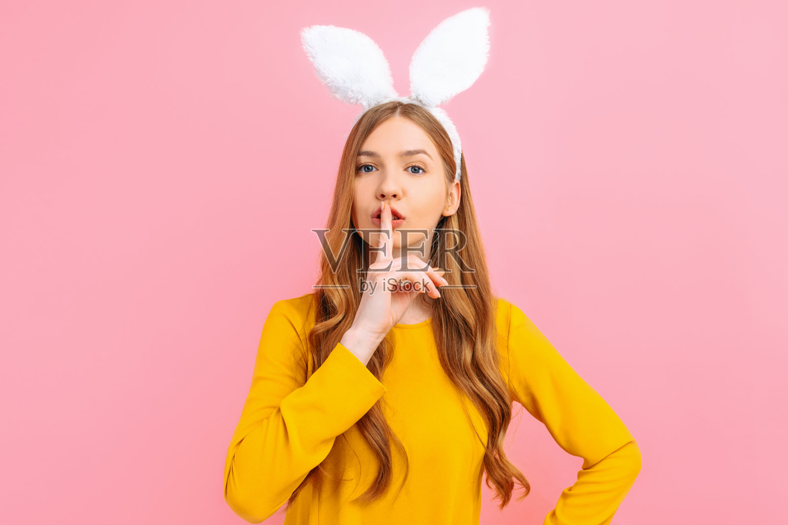 在粉红色的背景下，小女孩竖起复活节兔子的耳朵，用食指捂住嘴唇，做了一个安静的手势照片摄影图片