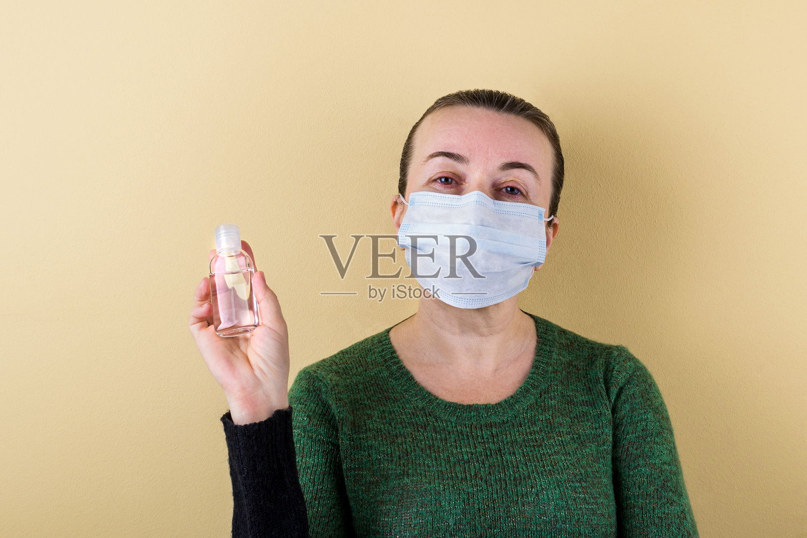 一名白人成年妇女穿着毛衣，脸上戴着医用口罩，手里拿着一瓶消毒凝胶。米色背景。照片摄影图片