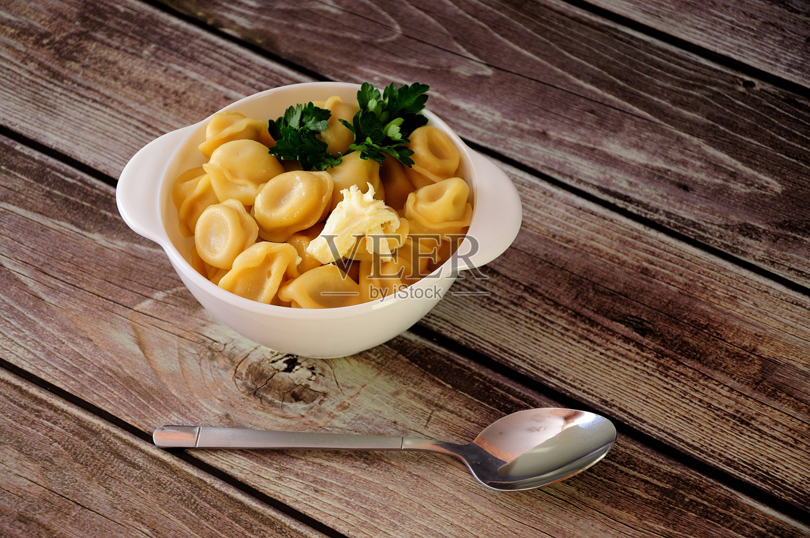 木桌上放着一个白色陶瓷盘子，上面放着刚煮好的饺子、一根欧芹和一片黄油。照片摄影图片
