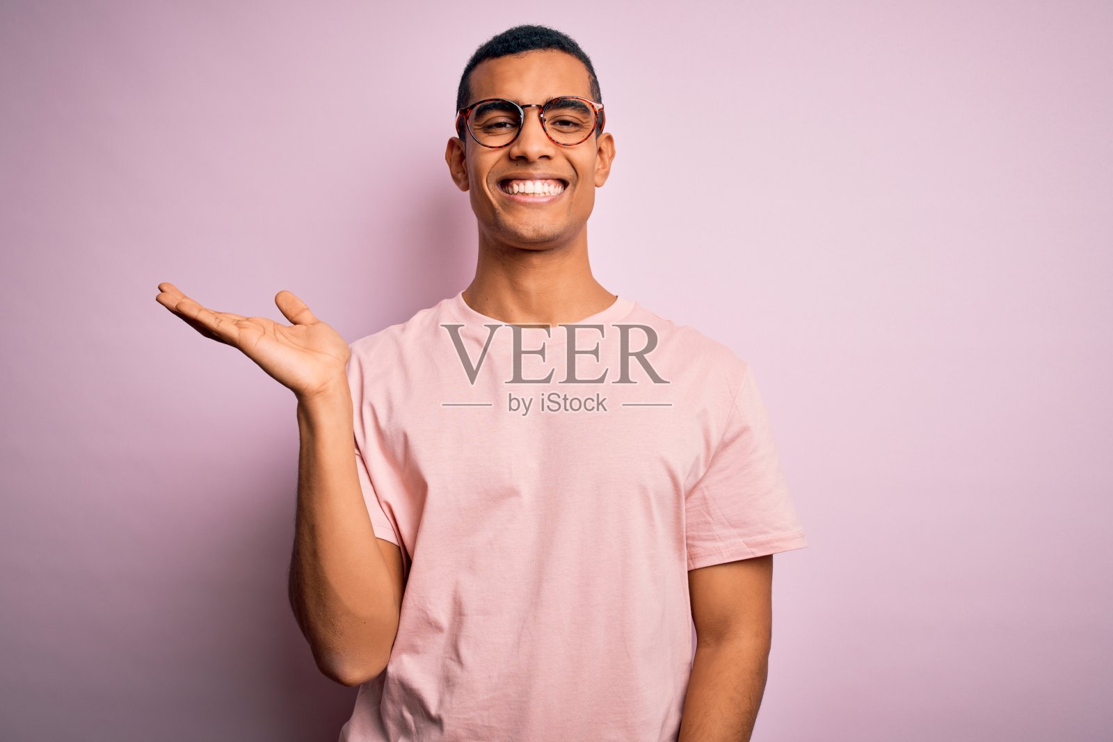 英俊的非裔美国人穿着休闲t恤和眼镜在粉红色的背景，微笑着提出和指向与手掌看着相机。照片摄影图片