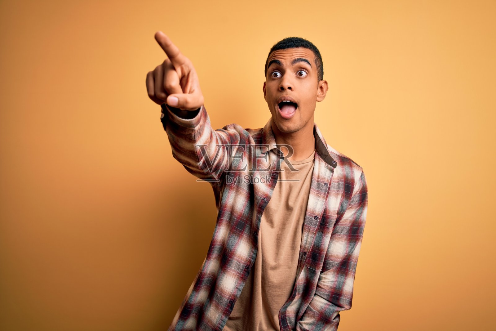 年轻英俊的非裔美国人穿着休闲衬衫站在黄色的背景上，用手指惊讶地指着前方，张着嘴惊讶地表情，前面有东西照片摄影图片