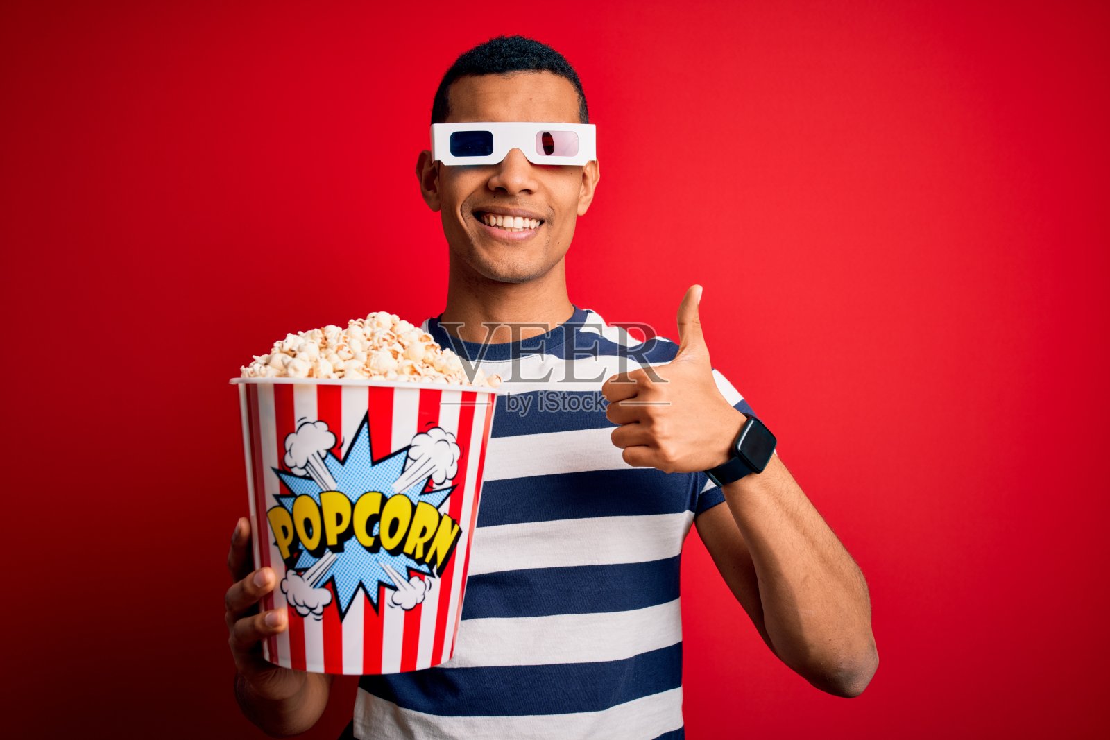 年轻英俊的非裔美国人看电影用3d眼镜吃爆米花做快乐的拇指手势与手。赞许的表情看着镜头，表示成功。照片摄影图片