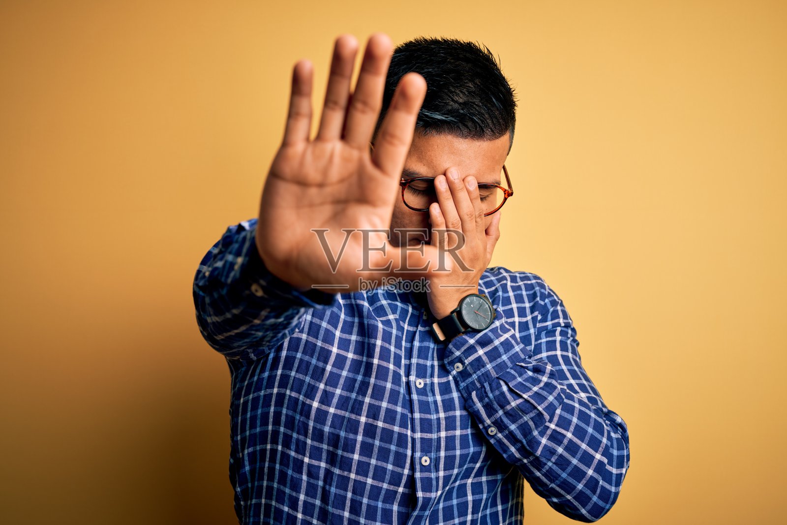 年轻英俊的拉丁男子穿着休闲衬衫和眼镜在黄色背景覆盖眼睛与手做停止手势与悲伤和恐惧的表情。尴尬和消极的概念。照片摄影图片
