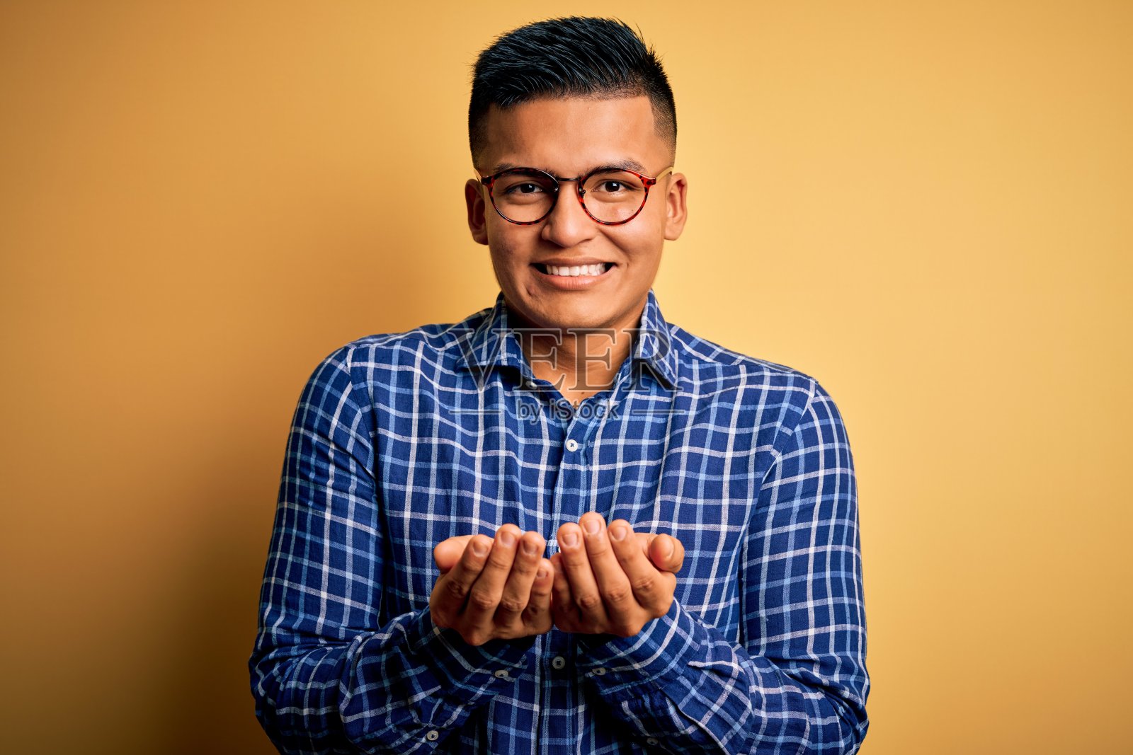 年轻英俊的拉丁男子穿着休闲衬衫和眼镜在黄色背景微笑与手掌在一起接受或给予的手势。保存和保护照片摄影图片