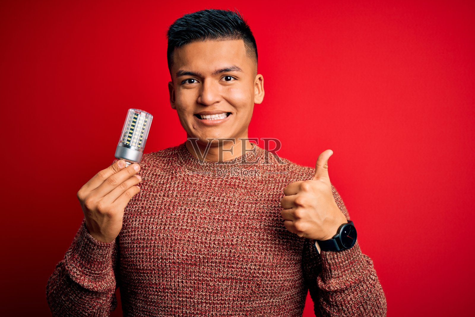 年轻英俊的拉丁男子在孤立的红色背景下举着led灯泡，开心地笑着做ok的手势，用手指竖起大拇指，优秀的手势照片摄影图片