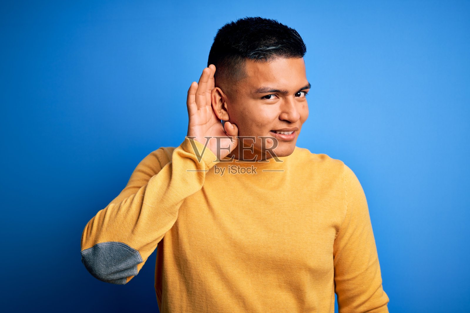 年轻英俊的拉丁男子穿着黄色休闲毛衣在孤立的蓝色背景微笑与手捂耳朵听谣言或流言。耳聋的概念。照片摄影图片