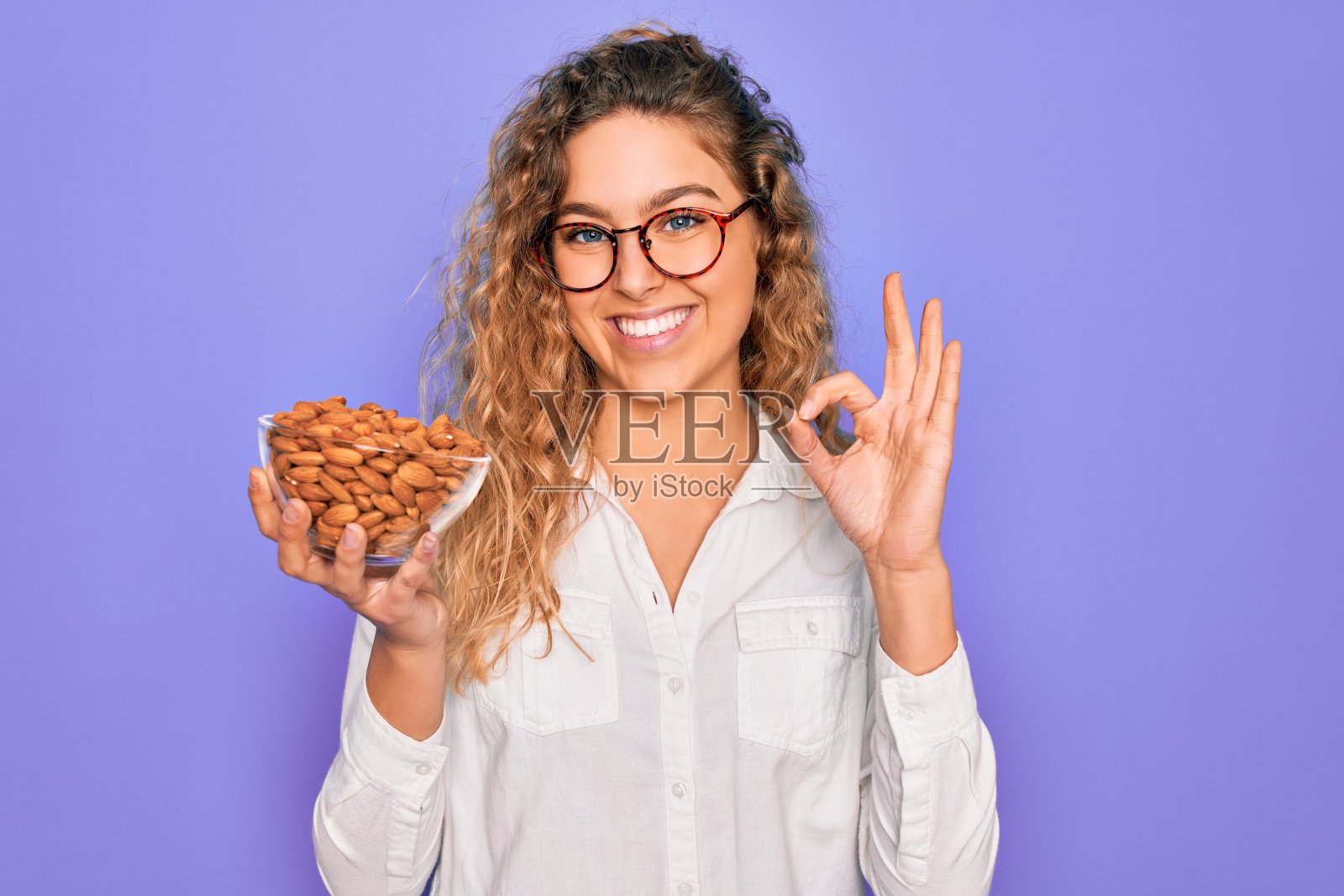 年轻美丽的金发碧眼女人拿着碗与健康的杏仁零食做ok手势与手指，优秀的象征照片摄影图片