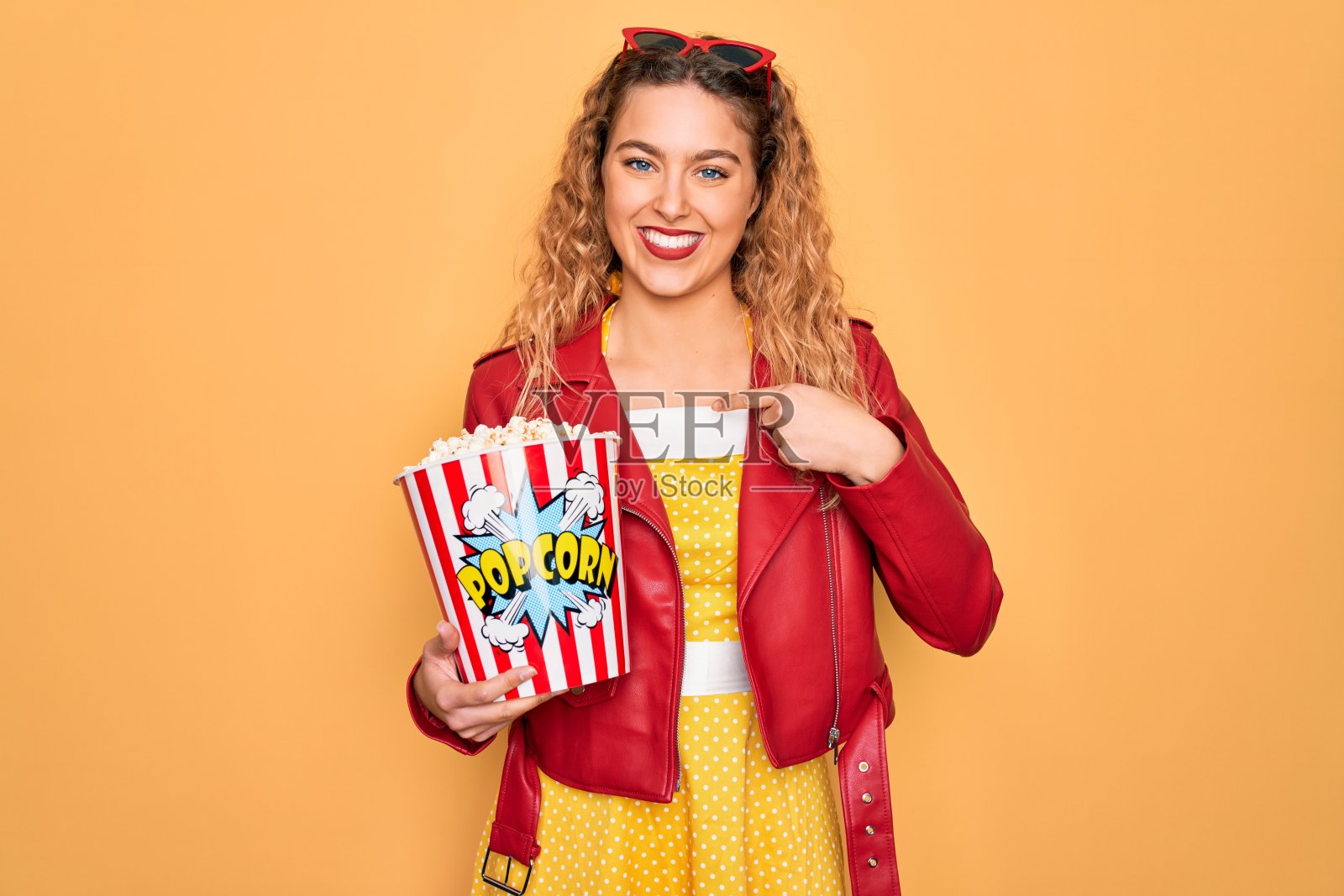 美丽的金发女人与蓝色眼睛吃咸爆米花零食在黄色的背景与惊讶的脸指向自己照片摄影图片