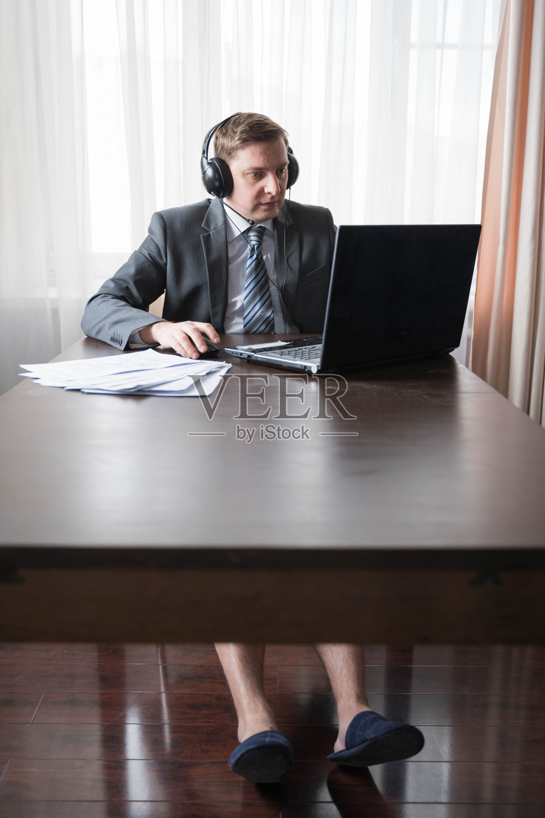 年轻的失业商人穿着灰色西装、衬衫和拖鞋在远程办公，因为冠状病毒大流行COVID-19的检疫。远程工作在家的桌子。通过笔记本电脑与老板进行耳机在线会议照片摄影图片