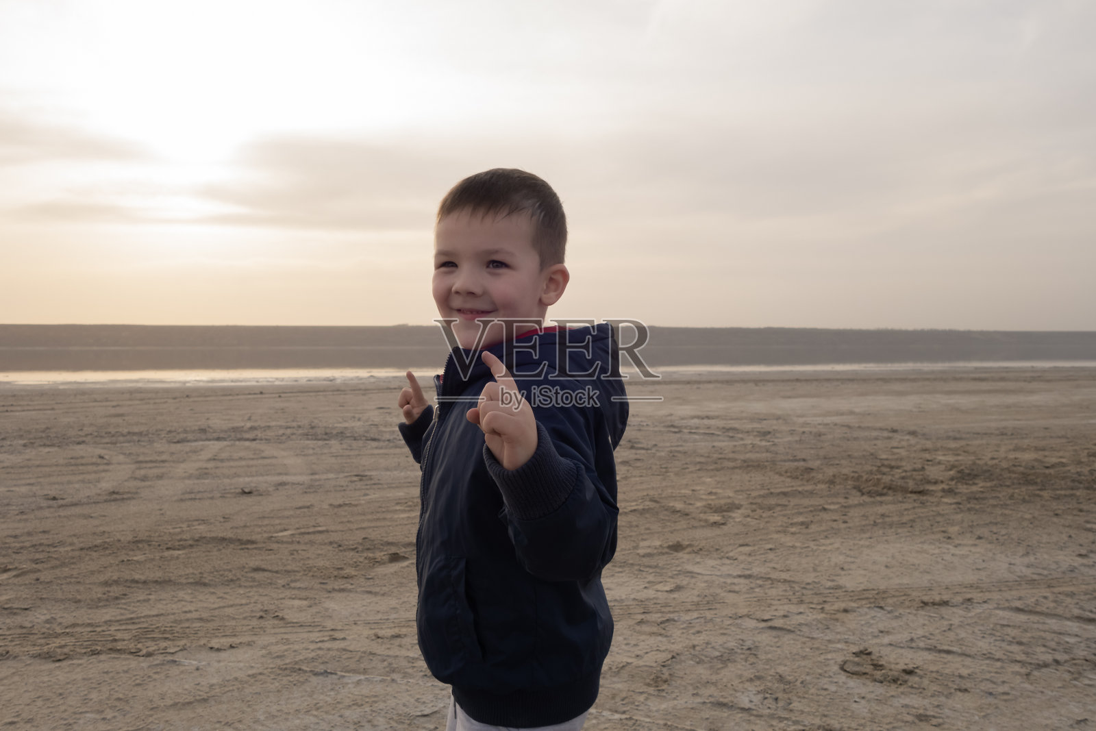 一个4岁穿着蓝色夹克的男孩在空旷的海滩上。描绘了一个超级英雄。照片摄影图片