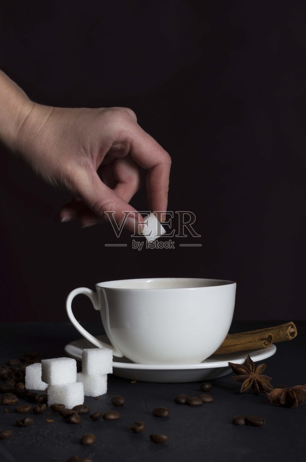 垂直拍摄。黑色的桌子上放着方糖，肉桂棒，咖啡豆，还有一个女人把方糖放进白色的杯子里。准备美味的热咖啡的过程照片摄影图片