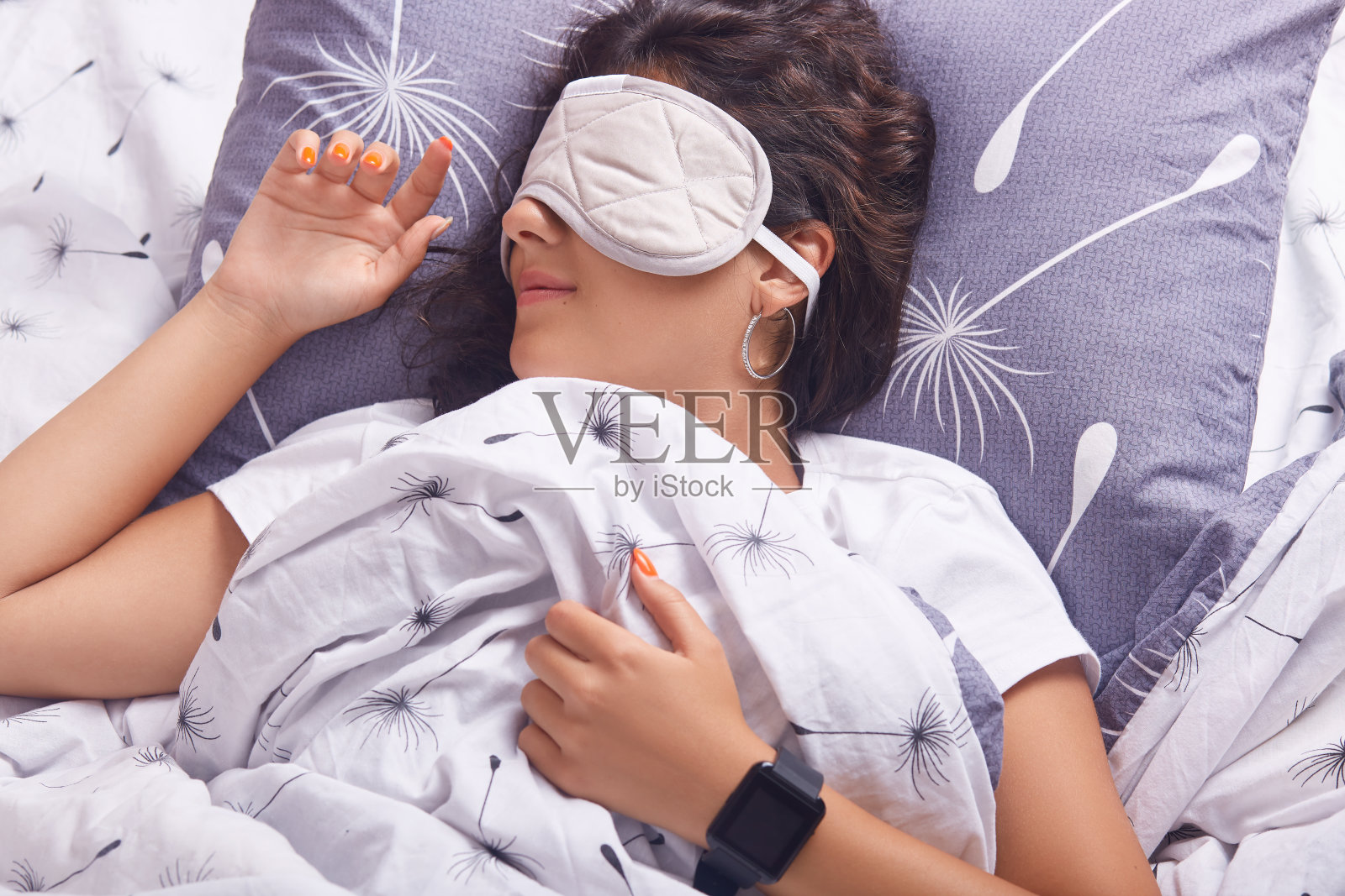 可爱的女孩用睡眠面具遮住眼睛，躺在床上的枕头上，一位不想早起的女士，一个裹着毯子的女人，手上戴着智能手表。照片摄影图片