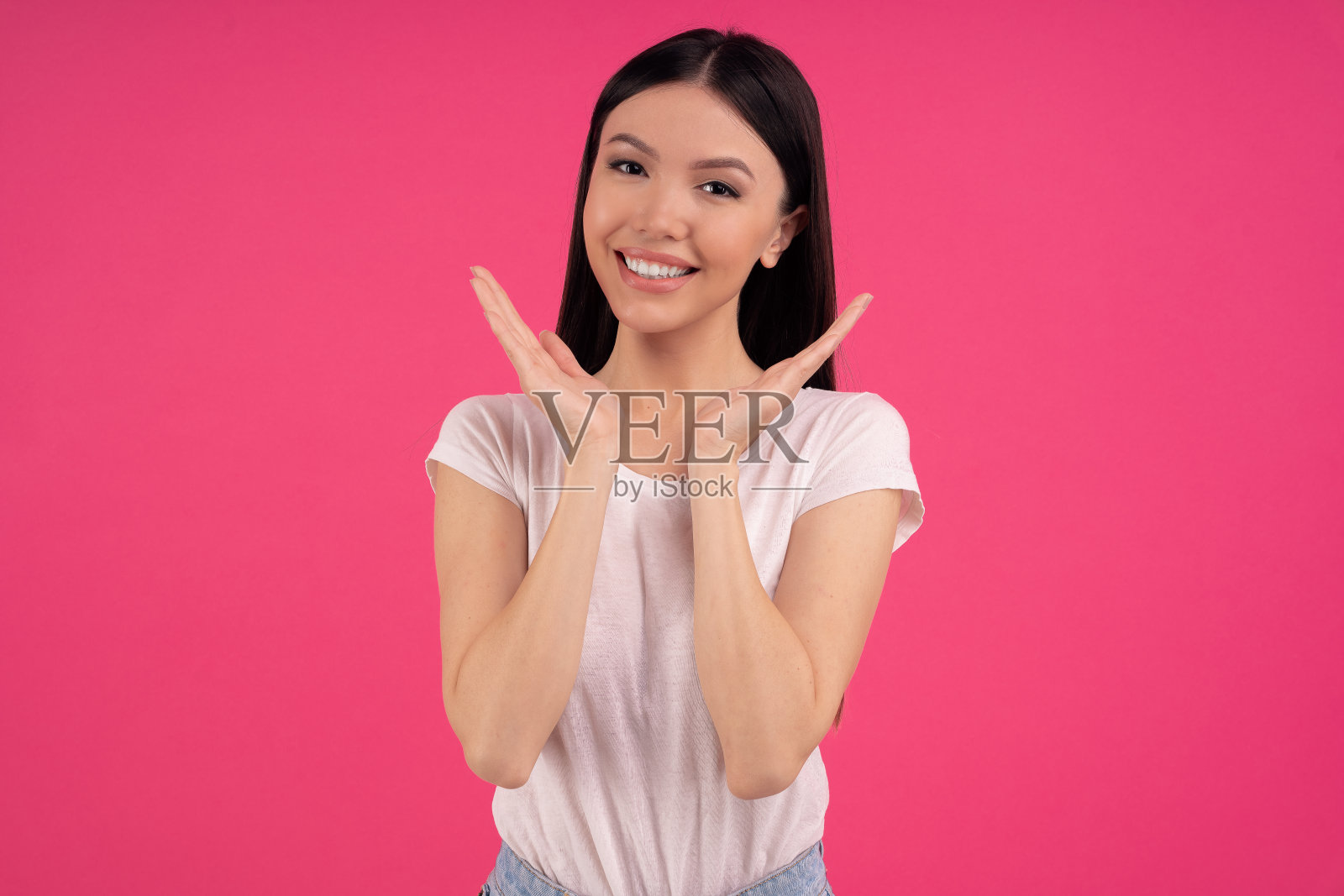 长相宜人的亚洲女模特开心地微笑，向脸附近摊开手掌，表达积极的情绪，穿着白色t恤，有吸引力的外表，健康的皮肤，隔离在粉红色的工作室墙壁上照片摄影图片