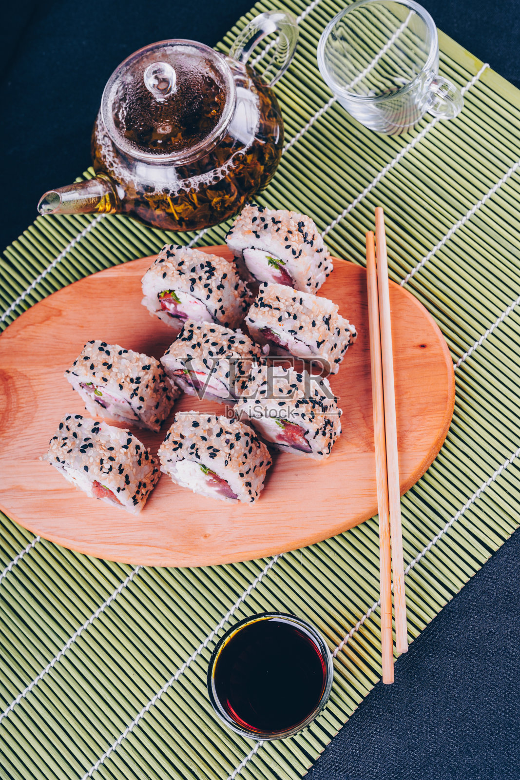美味的卷在木板上加上酱油和绿茶在装饰的竹餐巾上。近距离的美味寿司卷与筷子在砧板上的黑色背景照片摄影图片