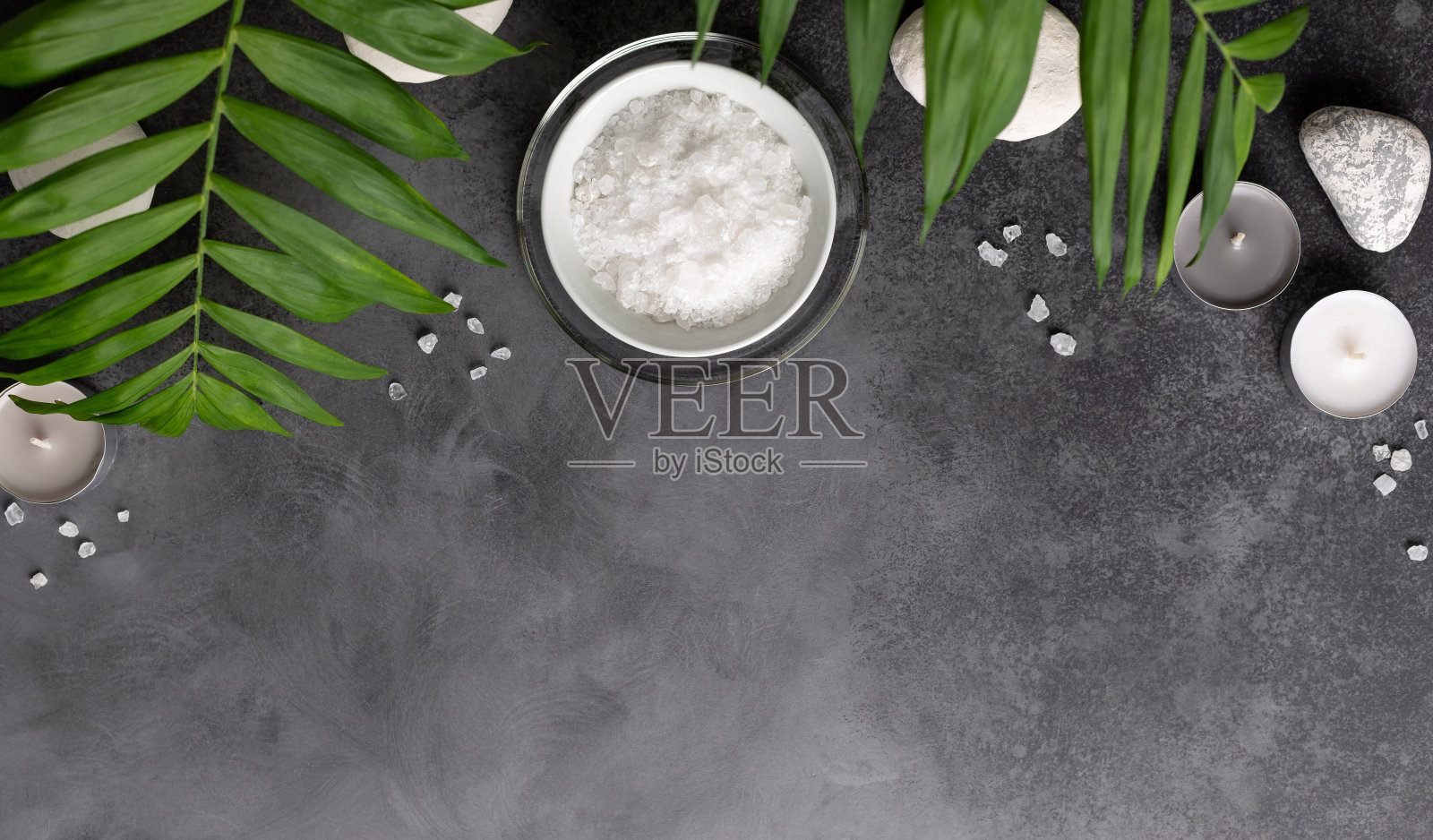 水疗设置与天然海盐碗，蜡烛和绿色棕榈叶在黑暗的石头背景。照片摄影图片