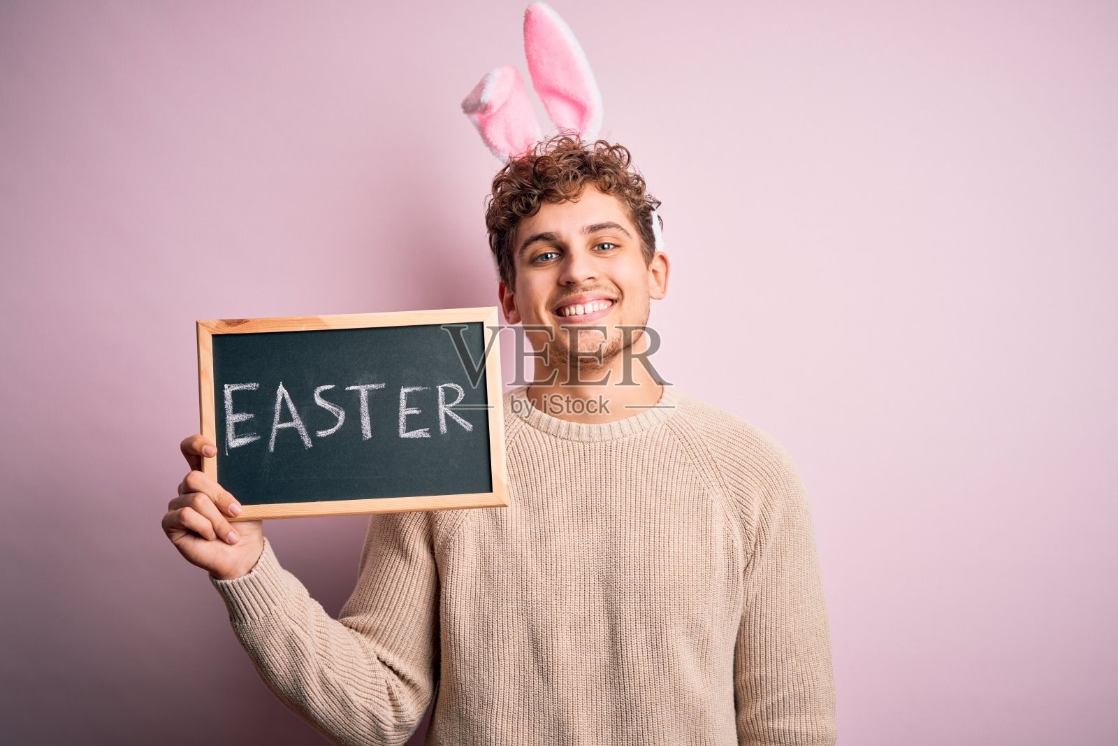 年轻英俊的金发男子戴着复活节兔子耳朵，拿着印有节日单词的黑板，带着快乐的脸站着，微笑着，自信地笑着露出牙齿照片摄影图片