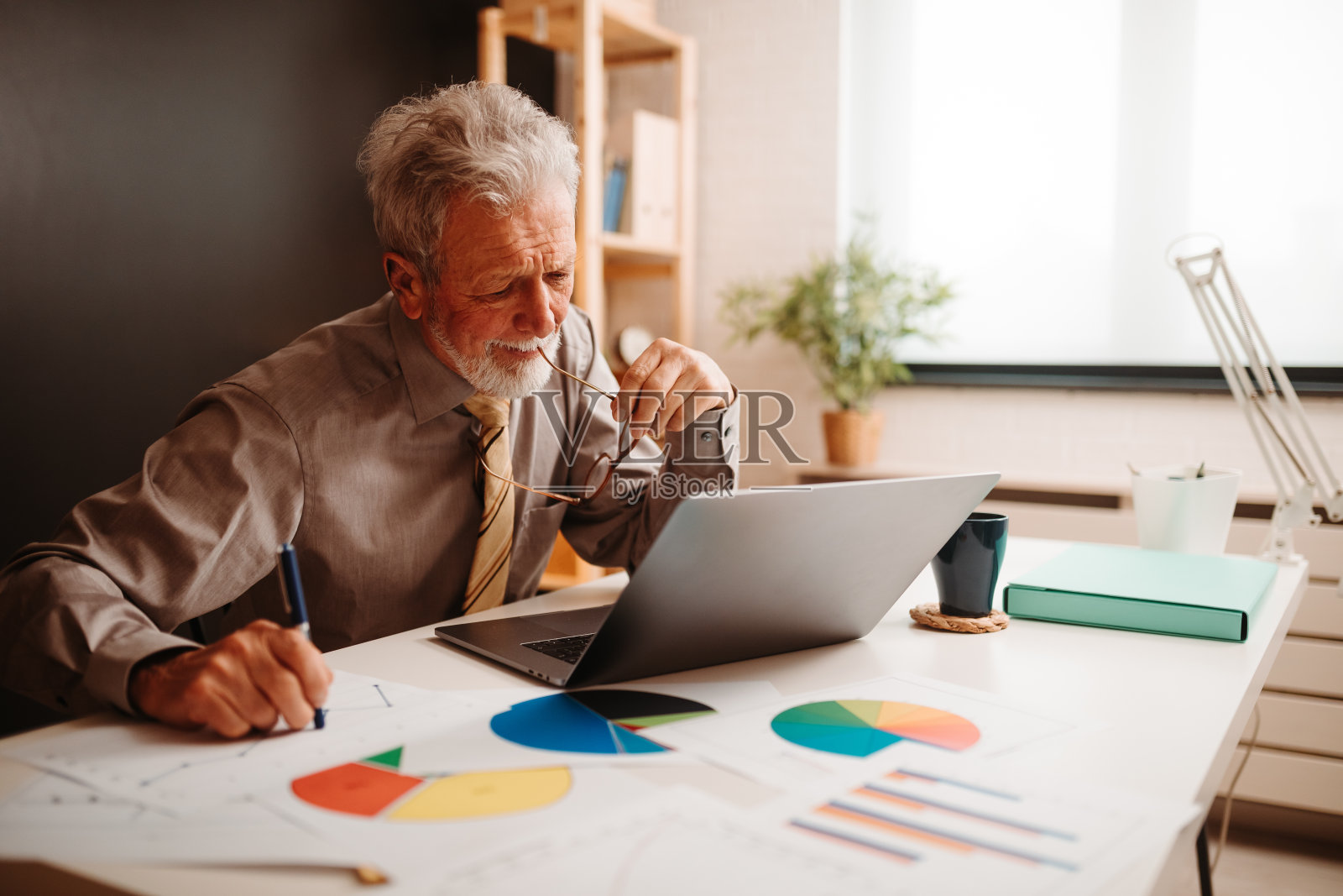 沮丧的商人坐在办公室里看笔记本电脑的肖像。他一只手拿着笔，另一只手拿着眼镜。照片摄影图片