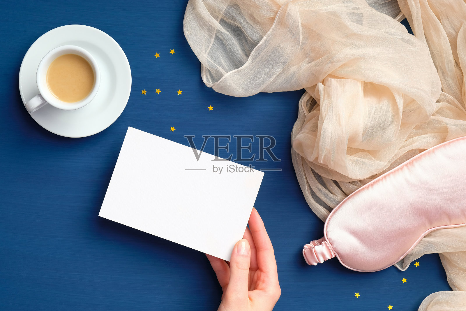 女性手握空白纸卡在经典的蓝色背景与咖啡杯和粉色睡眠眼罩。快乐清晨，早起，保持清醒的理念。平躺，俯视图照片摄影图片